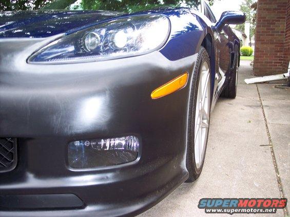 Blue Chevrolet Corvette front bumper with black unpainted section, headlight, and orange turn signal. Low angle