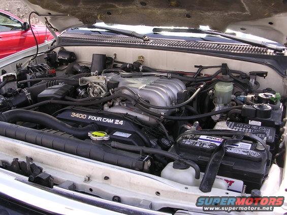 Engine bay with a V6 engine. Text on the engine reads '3400 FOUR CAM 24'. A red car is partially visible on the left