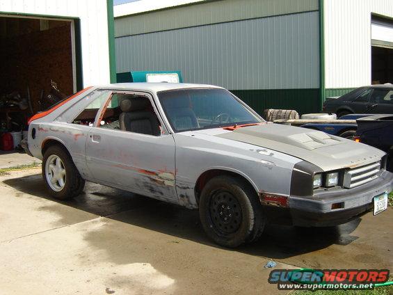 1983 Mercury Capri in primer with a hood scoop, parked outside a garage
