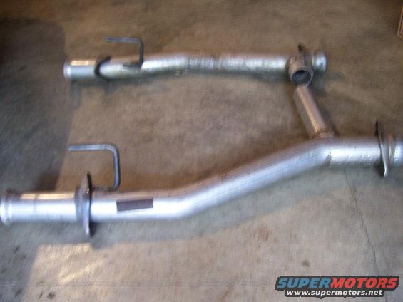 Two exhaust pipes, one longer and cleaner than the other, laid on a concrete floor. A dark rectangular sticker is visible on the longer pipe