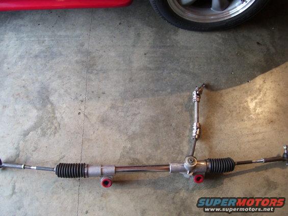 Steering rack with red mounting bushings and black bellows, on concrete floor with part of a red car and a silver wheel visible