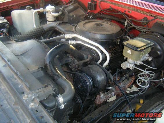 Engine bay with V8 engine, air cleaner, brake fluid reservoir, and hoses. Red exterior paint visible