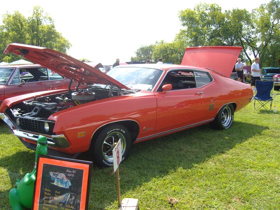 Orange 1970 Ford Torino GT, 3q-front view, hood and trunk open. Display sign visible