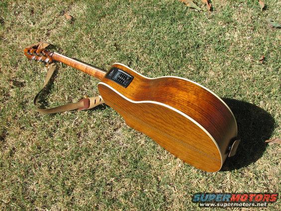 Wooden acoustic guitar with a strap lying on grass. The body is brown with white binding. A black electronic tuner is visible on the side