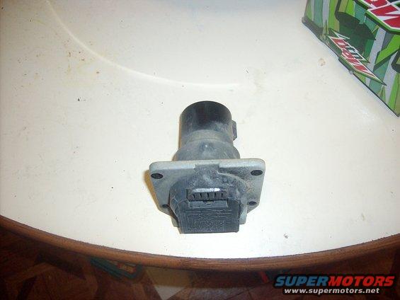 Trailer hitch connector with a black top and gray base, sitting on a table. A green and white box is in the background