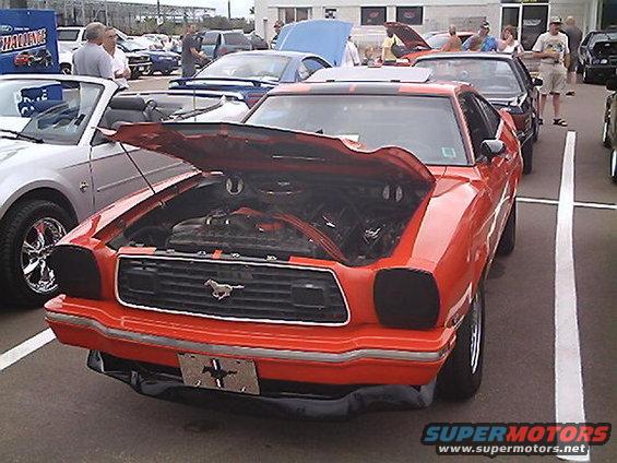 Orange 1978 Ford Mustang II, hood open, parked at car show. Other cars visible