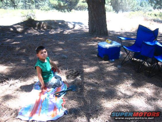 Child in green shirt sits on colorful blanket with shovel. Blue cooler and chairs nearby under tree