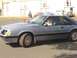 1986 Ford Mustang - A gray coupe with a sunroof is driven by a person, with other vehicles and a building in the background.