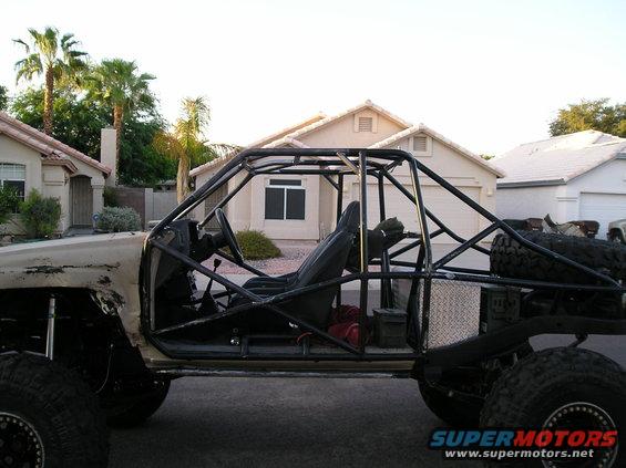 Tan off-road buggy with custom black roll cage, exposed engine bay, and large tires. Modified body panels show wear