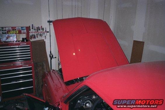 Red car interior with dashboard gauges visible, hood open, parked in a garage with tools