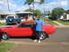 1967 Ford Mustang - A man poses next to a bright red 1967 Ford Mustang coupe on a sunny day, with other vehicles and palm trees in the background.