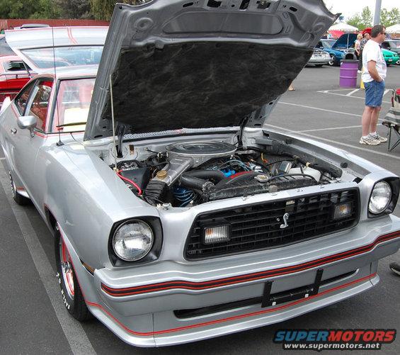 Silver 1970s coupe front view, hood open showing engine. Red pinstripes on body