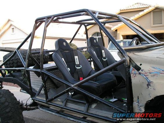 Off-road buggy interior with racing seats, harnesses, and a full roll cage. Spare tire visible