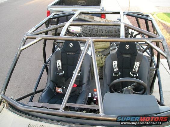 Custom off-road vehicle interior with roll cage, two black racing seats with Crow harnesses, and a spare tire visible behind the seats