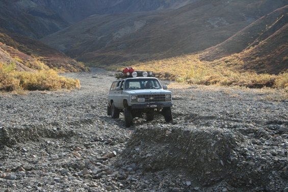 Gray Chevrolet Suburban with roof rack driving on a rocky, dry creek bed