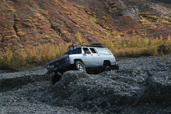 Silver 1990 Chevrolet Suburban driving over rocks with roof rack and cargo box