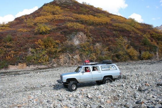 Silver 1990 Chevrolet Suburban driving on a rocky riverbed with a mountain in the background