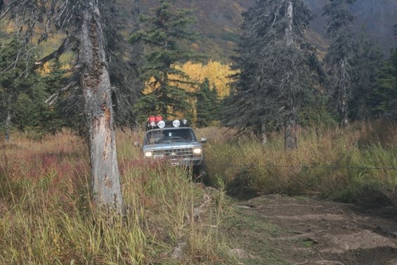 Silver Chevrolet Suburban with roof rack and lights driving on a dirt path through tall grass and trees