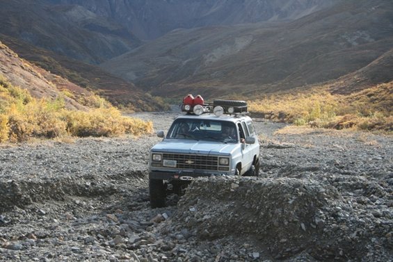 Light blue SUV with roof rack, gas cans, and spare tire driving through a rocky canyon