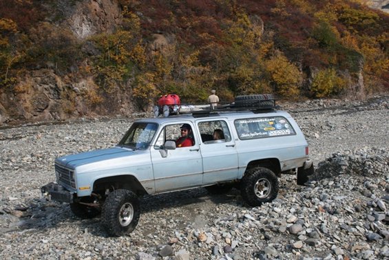 1990 Chevrolet Suburban with roof rack and spare tire, driving on a rocky riverbed