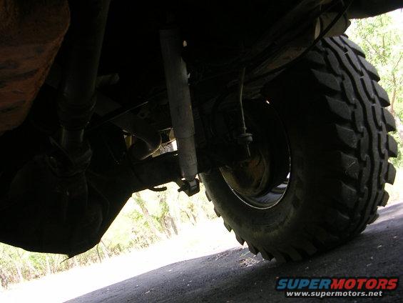 Undercarriage view of a vehicle's rear axle, shock absorber, and large tire. The tire has aggressive tread. Text 'SUPERMOTORS' is visible