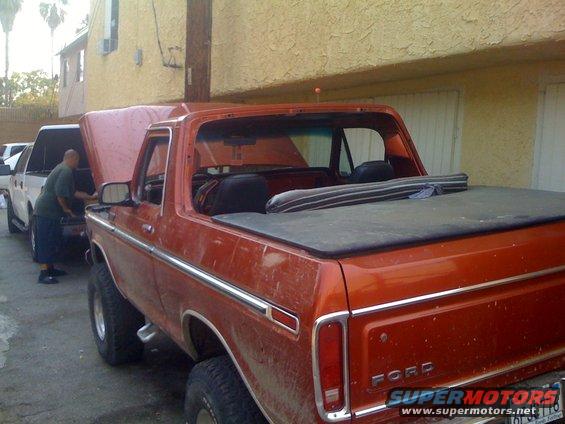 Orange 1978 Ford Bronco with a black tonneau cover and chrome trim, parked next to another vehicle with its hood open
