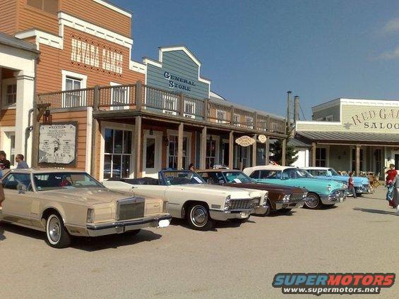 Several classic American cars, including a beige coupe and a white convertible, are parked in front of buildings designed to look like a Western town