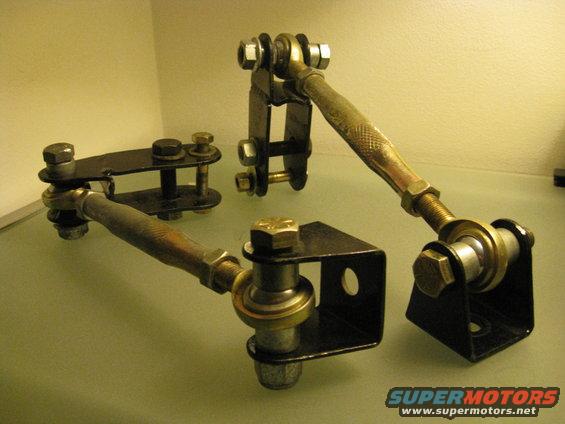 Two automotive suspension linkage parts with metal rods, ball joints, and mounting brackets on a reflective surface