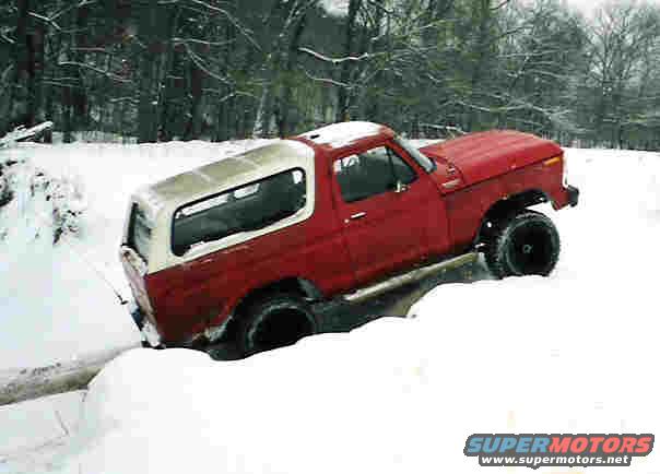 Red and white 1978 Ford Bronco partially submerged in a snow-filled ditch, with trees in the background
