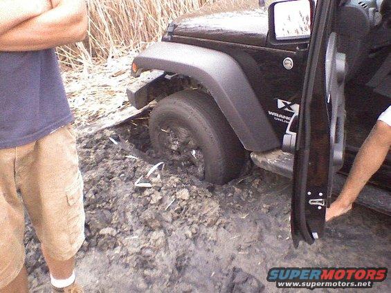 Black Jeep Wrangler stuck in mud, front wheel deep, door open, person standing nearby