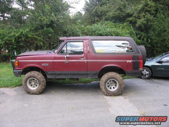 1990 Ford Bronco side profile, lifted with large tires. Another car is parked to the right