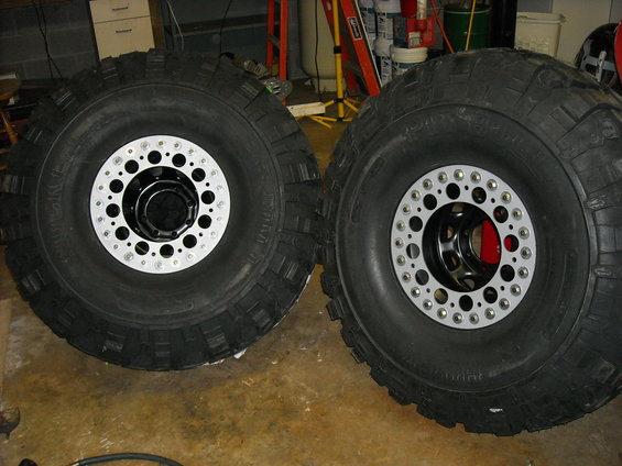 Two large off-road tires with custom multi-hole silver and black wheels are shown side-by-side on a concrete floor