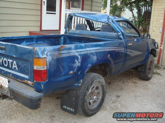 Blue Toyota pickup truck with dented roof and body damage, seen from the rear three-quarter angle