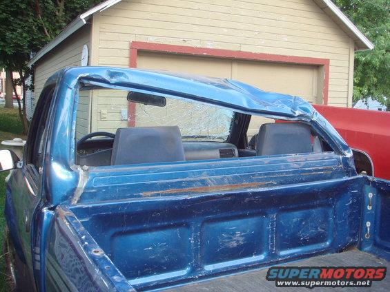 Blue pickup truck's roof is crushed inward, with shattered rear window. Red vehicle visible behind it