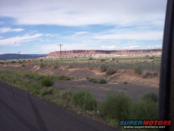 Distant white vehicles on a road next to a red rock formation under a cloudy sky