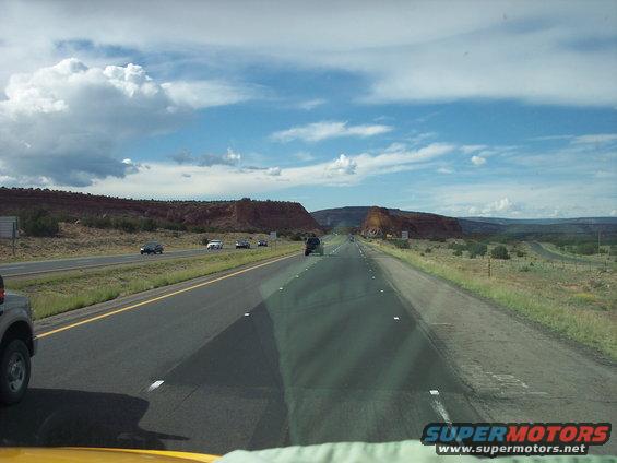 Highway driving through a desert landscape with red rock formations and scattered vehicles under a cloudy blue sky