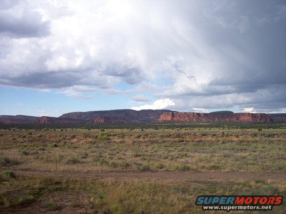 Wide shot of desert mesas under a cloudy sky, with dry grass and brush in the foreground