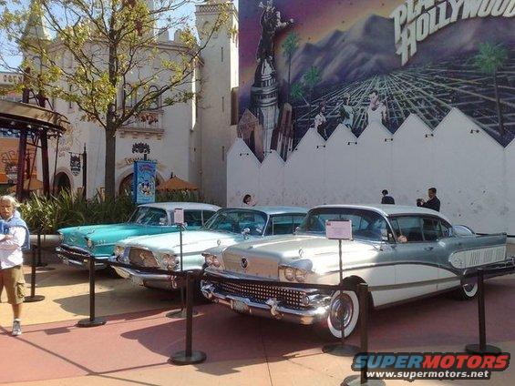 Three classic cars are lined up in front of a large mural depicting a cityscape and mountains. A person stands to the left