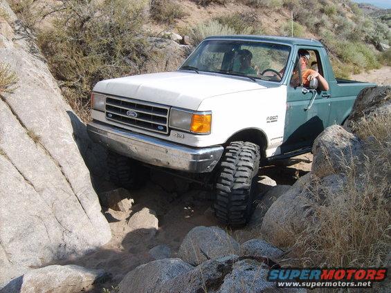 White and green Ford Bronco navigating rocky terrain. The front grille and headlights are visible