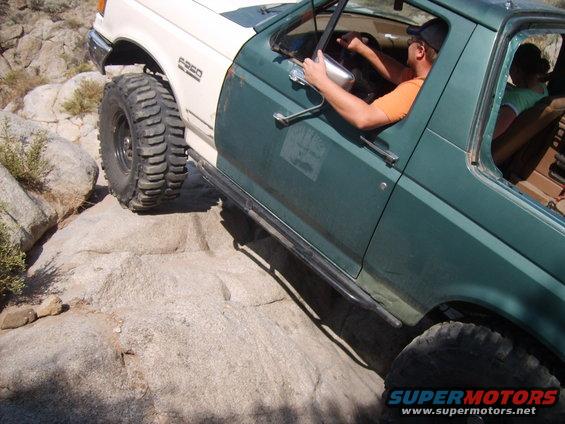 Green and white Ford F-250 with large tires navigating rocks. Driver visible