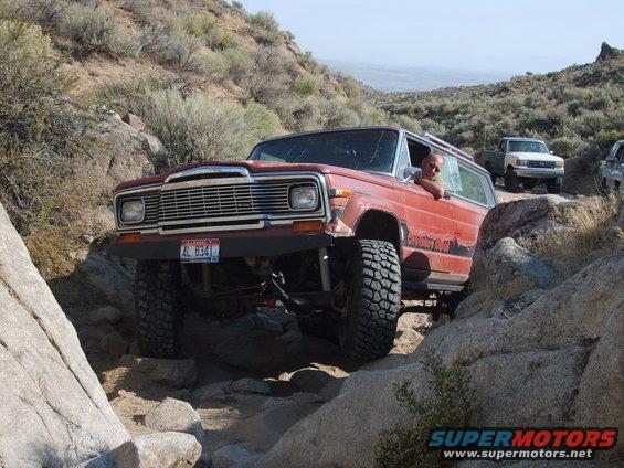 Red Jeep Wagoneer off-roading on rocks, driver visible, other vehicles behind