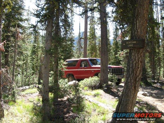 Red Ford Bronco parked on a dirt path in a forest, with a sign pointing to Seafoam Lake