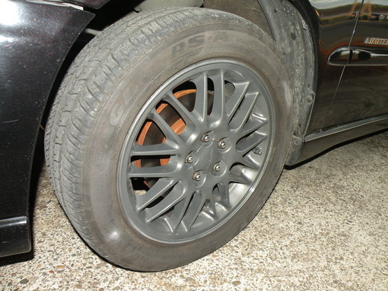 Close-up of a car's front wheel and tire with a grey multi-spoke rim and orange brake rotor