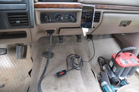 Dashboard and floor of a vehicle, showing pedals, a phone mount with a phone, various cords, a diagnostic tool, and a car battery with tools