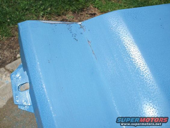 Close-up of a blue painted metal panel with some rust spots and a crack. A mounting bracket is visible on the left