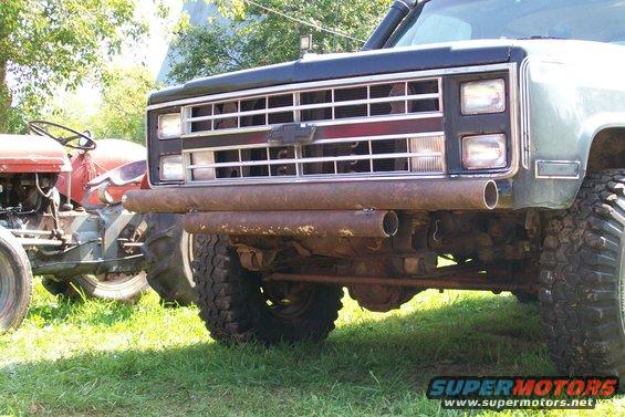 Front end of a dark SUV with a chrome grille and a rusty pipe bumper. A red tractor is parked next to it