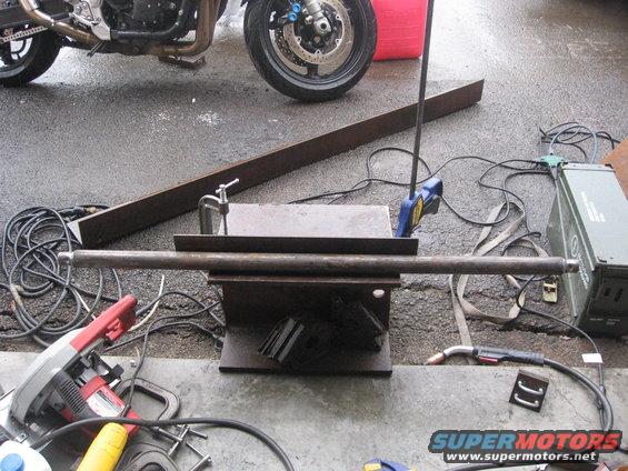 Motorcycle parked near metal pipe clamped in vise, with welding gear and saw nearby