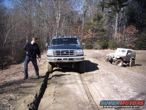 Blue Ford F-Series truck stuck in mud with a white Jeep Wrangler in the background also stuck