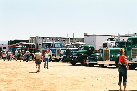Several trucks, including green and blue models, parked outdoors with people milling around