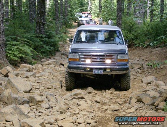 1996 Ford Bronco driving on a rocky, wooded trail with other vehicles behind it